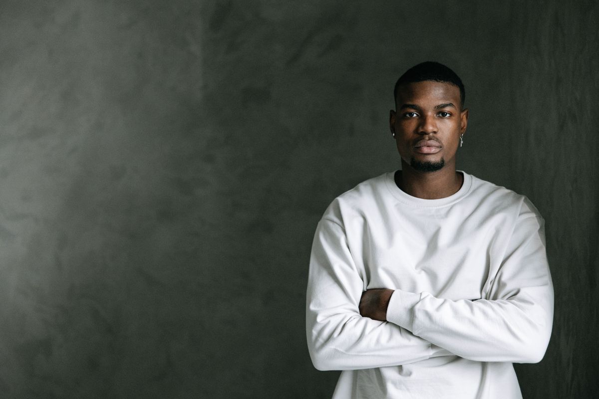 A young man in a white sweater, arms folded, steady gaze, against a soft grey studio wall